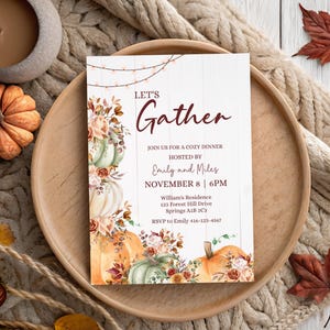 May include: A rectangular invitation card with a white wood-plank background featuring string lights at the top and watercolour pumpkins and autumn flowers along the sides. The text reads: LET'S Gather, JOIN US FOR A COZY DINNER HOSTED BY Emily and Miles, NOVEMBER 8 | 6PM, William's Residence, 123 Forest Hill Drive, Springs A1B 2C3, RSVP to Emily 416-123-4567.