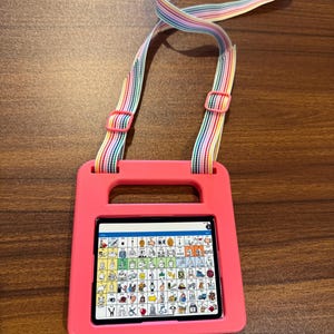 May include: A tablet device encased in a bright pink protective frame with a handle, featuring a rainbow-striped adjustable carrying strap. The screen displays a grid of colorful communication icons and symbols used for augmentative speech.