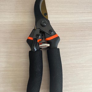 May include: Black and orange pruning shears with a curved blade and padded black handles. The shears have a gold-colored blade and are designed for trimming plants and shrubs. The tool is shown on a light wood surface.