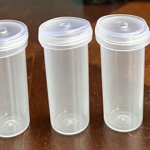 May include: Five clear plastic vials with white lids are arranged in a row. The vials are cylindrical with a slightly angled top. The lids have a small, raised center. The vials are empty and appear to be new.