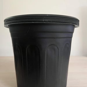 May include: A black plastic plant pot with a ridged rim and vertical arched indentations. The pot is cylindrical and sits on a light-colored surface. The pot is empty and ready for planting.