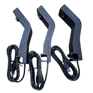 May include: Three black, textured, curved stands with attached black USB cables. Each stand has a curved arm and a base with a clip. The cables are neatly bundled. The stands are designed for electronic devices.