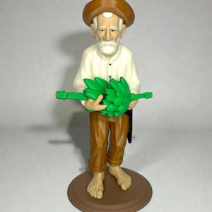 May include: A figurine of an elderly man wearing a brown hat, white shirt, and brown pants, holding a green object. The figure stands barefoot on a brown circular base. The figurine has a white beard and mustache.