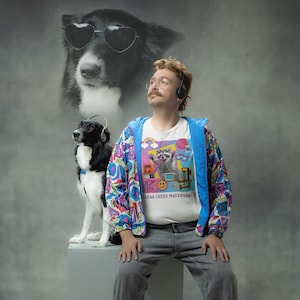 May include: A man wearing a white t-shirt with a raccoon graphic and the text "CLEAR CREEK MAKERSPEACE", a colorful jacket, and gray pants. A dog wearing headphones sits next to him. A dog wearing heart-shaped sunglasses is in the background.