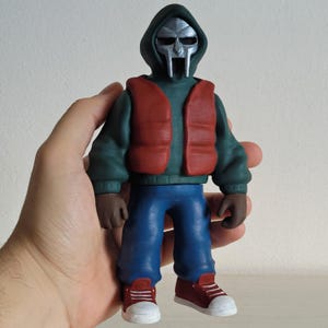 Puede incluir: Figura de un personaje con máscara plateada, sudadera con capucha verde, chaleco rojo, pantalones azules, guantes marrones y zapatillas rojas. La figura está sujeta con una mano.