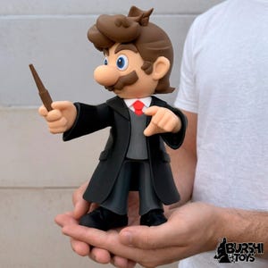 Puede incluir: Una figura de un personaje con cabello castaño y bigote, vistiendo un abrigo negro y sosteniendo una varita. El personaje tiene ojos azules, una corbata roja y un chaleco gris. La figura es una mezcla de Mario y Harry Potter.