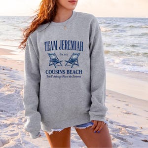 May include: A gray sweatshirt with the text "TEAM JEREMIAH" above "COUSINS BEACH" and the phrase "We'll Always Have the Summer". The design includes two blue beach chairs and the year "Est 2022".