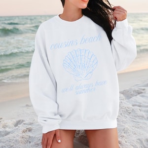 Puede incluir: Sudadera blanca con el texto "cousins beach" y "we'll always have summer" en azul claro. Un gráfico de concha marina azul claro está debajo. Una prenda informal con temática playera.