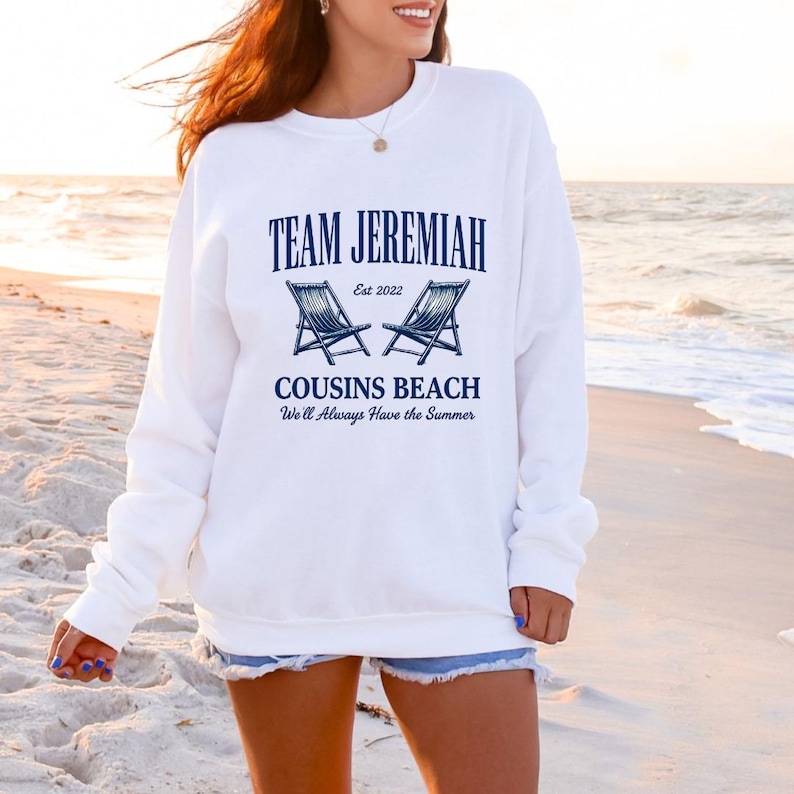 May include: White sweatshirt with the text "TEAM JEREMIAH" above "COUSINS BEACH" and the phrase "We'll Always Have the Summer." The design includes two blue beach chairs. The sweatshirt is worn on a beach.