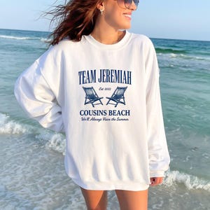 May include: White sweatshirt with navy blue text that reads "TEAM JEREMIAH Est 2022" above two blue beach chairs. Below the chairs, the text reads "COUSINS BEACH We'll Always Have the Summer". The sweatshirt is worn by a person at the beach.