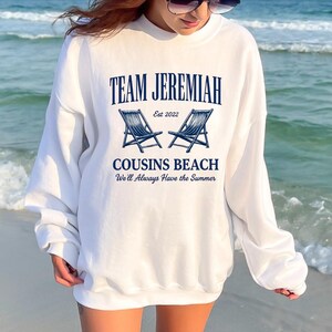 May include: White sweatshirt with the text "TEAM JEREMIAH" above two blue beach chairs and the words "COUSINS BEACH" and "We'll Always Have the Summer". The sweatshirt is worn on a beach.
