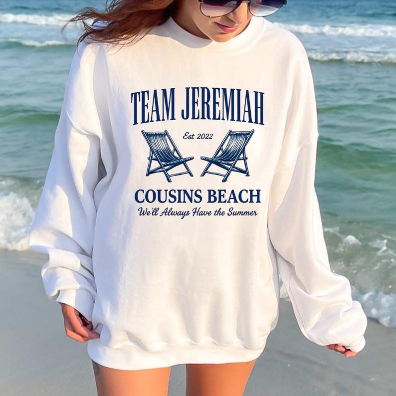 May include: White sweatshirt with the text "TEAM JEREMIAH" above two blue beach chairs and the words "COUSINS BEACH" and "We'll Always Have the Summer". The sweatshirt is worn on a beach.
