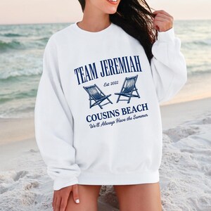 May include: White sweatshirt with the text "TEAM JEREMIAH" in navy blue, along with "COUSINS BEACH" and the phrase "We'll Always Have the Summer." Two navy blue beach chairs are also featured. The sweatshirt is a casual beach-themed apparel.