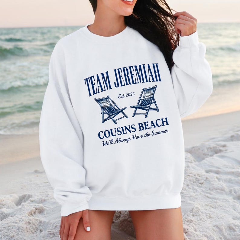 May include: White sweatshirt with the text "TEAM JEREMIAH" in navy blue, along with "COUSINS BEACH" and the phrase "We'll Always Have the Summer." Two navy blue beach chairs are also featured. The sweatshirt is a casual beach-themed apparel.