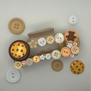 Pode incluir: Uma presilha de cabelo bege decorada com vários botões em tons de castanho, bege e branco. A presilha apresenta um botão de biscoito e um botão de urso de pelúcia. Botões adicionais estão espalhados ao redor da presilha.