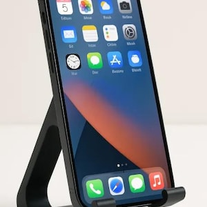 May include: A black smartphone is displayed on a black phone stand. The phone screen shows app icons and a blue and orange background. The stand has a rectangular base and a curved support.