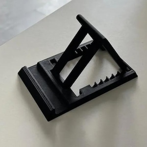 May include: A black, 3D-printed phone stand. The stand has a rectangular base with a triangular support structure and a notched edge to hold a phone. The stand is designed to hold a phone in a vertical position.