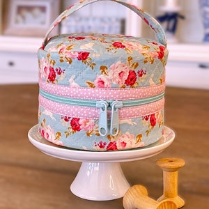 Pode incluir: Uma caixa de costura redonda e acolchoada com um padrão floral em rosa e vermelho sobre um fundo azul claro. A caixa tem um fecho de correr e uma pega azul claro. Está sobre um suporte branco para bolos, com bobinas de madeira e tesouras por perto.