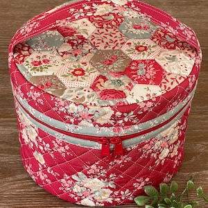 May include: A round, quilted cosmetic bag with a red floral pattern. The bag features a red zipper, a light blue accent stripe, and a matching red handle. The bag is approximately 8 inches in diameter and 6 inches tall.