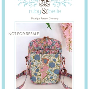 May include: A rectangular crossbody bag featuring a floral print with pink, blue, and green tones. The front panel displays a central illustration of a figure surrounded by blue flowers and foliage. It includes a ruffled pocket, rose gold hardware, and a matching fabric strap. The text 'NOT FOR RESALE' and 'Ava Cross Body Bag' are visible.