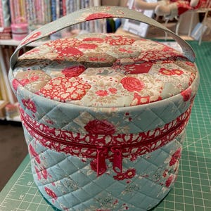 May include: A round, quilted fabric sewing box with a handle. The box features a floral pattern with red roses on a light blue background. A red zipper runs around the middle. The handle and top are a matching light blue fabric.