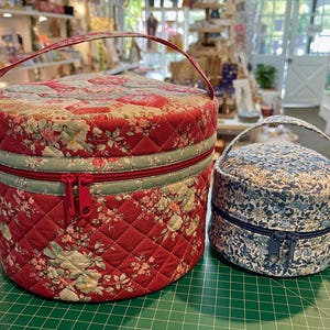 May include: Two round quilted fabric cases with handles. The larger case is red with a floral pattern and a red zipper. The smaller case is blue with a floral pattern and a blue zipper. Both cases have a quilted design.