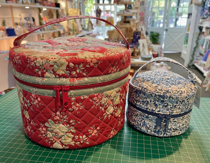 May include: Two round quilted fabric cases with handles. The larger case is red with a floral pattern and a red zipper. The smaller case is blue with a floral pattern and a blue zipper. Both cases have a quilted design.
