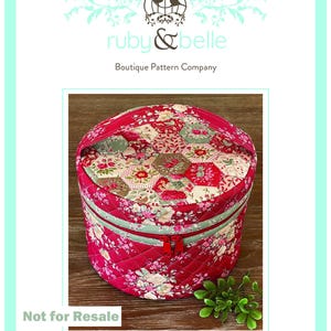 May include: A round, quilted train case with a red floral pattern and a mint green zipper. The case has a handle and a red zipper closure. The text "Rosalie Train Case" is at the bottom. The words "ruby & belle" and "Boutique Pattern Company" are at the top.