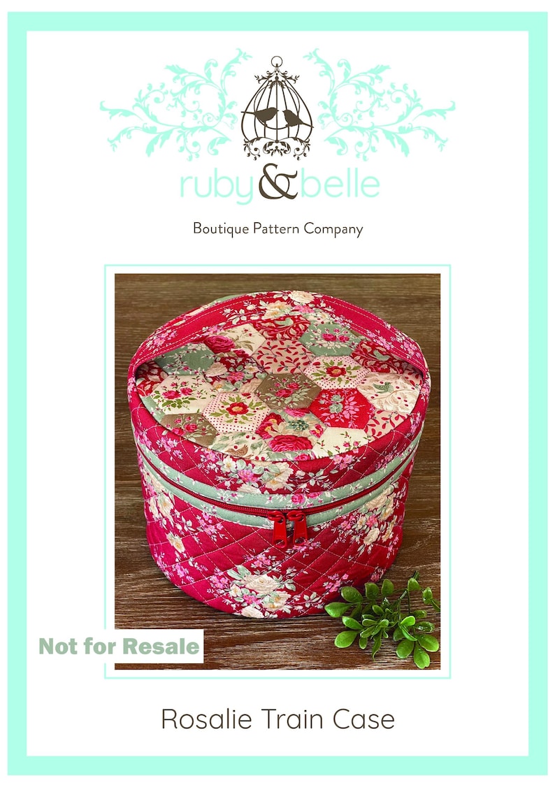 May include: A round, quilted train case with a red floral pattern and a mint green zipper. The case has a handle and a red zipper closure. The text "Rosalie Train Case" is at the bottom. The words "ruby & belle" and "Boutique Pattern Company" are at the top.