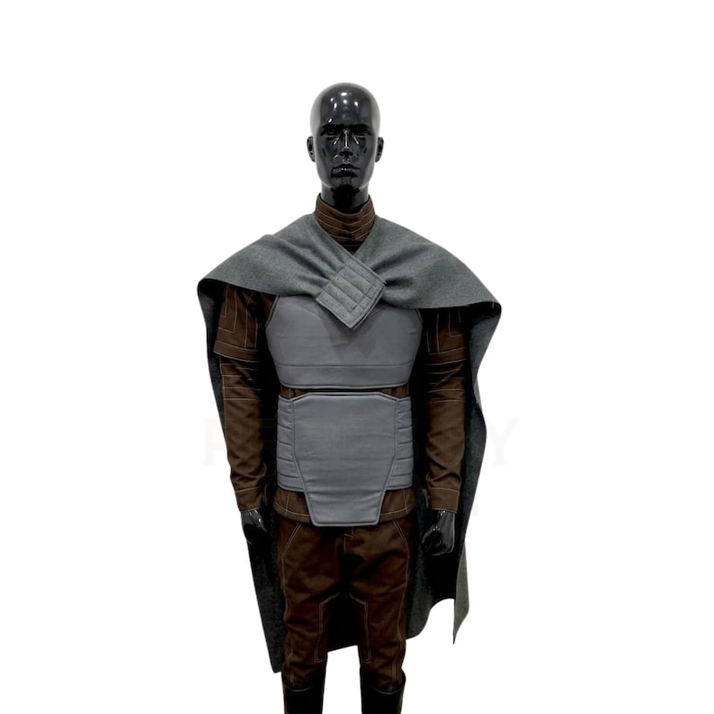 Pode incluir: Um traje completo exibido em um manequim preto. O traje inclui uma capa cinza, um protetor cinza para o peito e abd&ocirc;men, uma camisa marrom de manga comprida e cal&ccedil;as marrons. O traje tem um design futurista.