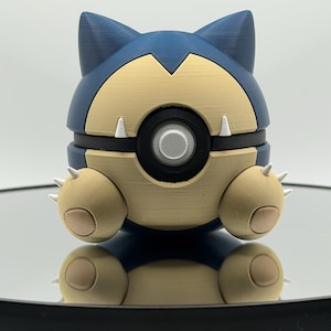 May include: A 3D-printed Snorlax figure in a sitting pose. The figure features a blue head and body with a tan belly and limbs. It has white claws and a black circular detail in the center. The figure is resting on a reflective surface.