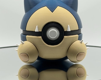 May include: A 3D-printed Snorlax figure in a sitting pose. The figure features a blue head and body with a tan belly and limbs. It has white claws and a black circular detail in the center. The figure is resting on a reflective surface.