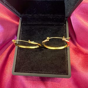 Pu&ograve; includere: Un paio di orecchini a cerchio ovali dorati, presentati in un cofanetto nero aperto. Gli orecchini hanno un design semplice e lucido. L'interno della scatola &egrave; in velluto nero, in contrasto con l'oro degli orecchini.