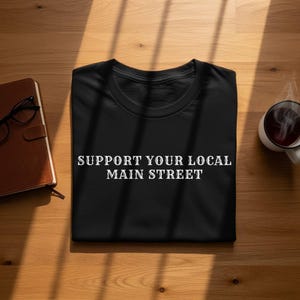 May include: A black short-sleeved t-shirt featuring the white text "SUPPORT YOUR LOCAL MAIN STREET" printed in a bold, serif font across the center of the chest. The garment is folded neatly, showcasing a classic crew neckline and a smooth fabric texture.