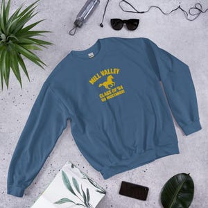 May include: A blue crew neck sweatshirt featuring the text "Mill Valley Class of '84 Go Mustangs!" in yellow, with a yellow horse graphic. The sweatshirt is displayed on a light grey surface.