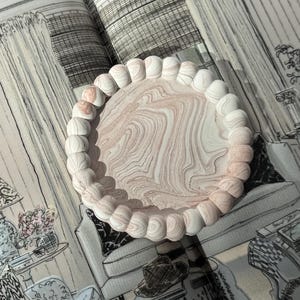 May include: A decorative tray with a light pink and white marbled design. The tray has a beaded edge and is set on a page with a black and white interior design illustration. The tray is a stylish home decor item.