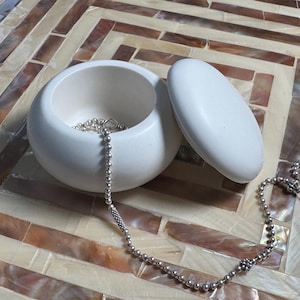 May include: A white, round jewelry box with a matching lid, next to a silver beaded necklace. The necklace is draped over the box and lid. The jewelry box is on a decorative surface with a mosaic pattern.