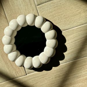 May include: A round, white decorative mirror with a unique, textured frame. The frame is made of rounded, bulbous shapes. The mirror is set against a light-colored wooden surface, with shadows cast across the wood.