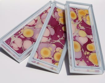 May include: Three rectangular trays with a marbled design in shades of pink, yellow, and purple. The trays have a light blue frame and are arranged at an angle, showcasing the unique patterns.