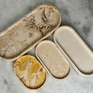 May include: A collection of four oval decorative trays in varying sizes. The trays feature a marbled design in shades of beige, brown, and yellow. The trays are made of a solid material and have a smooth, glossy finish. The trays are ideal for displaying jewelry or other small items.