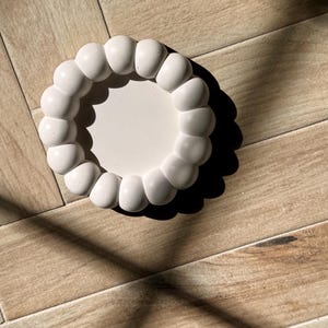 May include: A white, round decorative tray with a unique design. The tray features a series of rounded, bubble-like shapes around the edge, creating a textured border. The tray is sitting on a wooden surface.