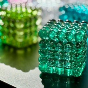 Puede incluir: Primer plano de un objeto decorativo en forma de cubo, construido con numerosas esferas verdes transparentes. Las esferas están dispuestas en un patrón de rejilla, creando una superficie texturizada. Otros objetos similares en verde y azul están borrosos en el fondo.