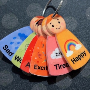 May include: A set of emotion keyrings with cartoon faces and labels. Emotions include Sad, Worried, Excited, Tired, and Happy. The Happy keyring features a rainbow. Keyrings are attached to a metal ring.