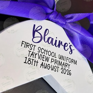 May include: A white wooden clothes hanger featuring a vibrant purple satin ribbon tied in a bow at the top. The hanger is printed with the text: Blaire's FIRST SCHOOL UNIFORM TAY VIEW PRIMARY 18TH AUGUST 2026 in a mix of purple and black lettering.
