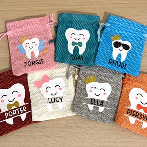 May include: Seven small, colourful burlap drawstring bags, each with a smiling tooth graphic and a name like "Jorgie" or "Rhudi". The bags are in pink, teal, blue, red, beige, grey, and orange. Ideal for small gifts or treats.