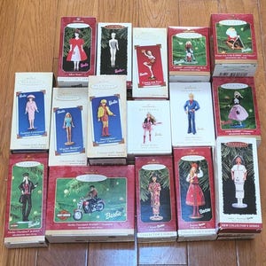 May include: A collection of Hallmark Keepsake Barbie ornaments in their original boxes. The boxes feature images of Barbie in various outfits and themes, including a Harley-Davidson Barbie and a Chinese Barbie. The boxes are red and green.