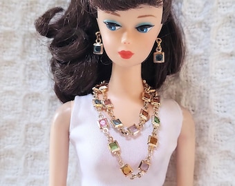 May include: A vintage doll with brown hair and blue eyes, wearing a white top and gold-toned jewelry. The jewelry includes a necklace and earrings, each featuring colorful square-cut gemstones. The doll's makeup includes red lipstick.