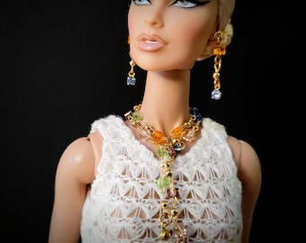 May include: A doll wearing a white crocheted top and a gold necklace with colorful beads. The doll also has matching earrings with colorful beads. The doll has blonde hair and is set against a black background.