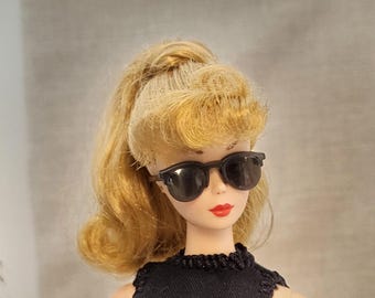May include: A vintage doll with blonde hair styled in a ponytail, wearing black sunglasses and a black top. The doll has red lipstick and a neutral background.