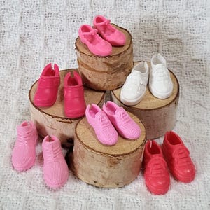 May include: A collection of miniature doll shoes in various styles and shades of pink and white. The assortment includes sneakers, Mary Janes, and clogs, arranged on small wooden platforms.